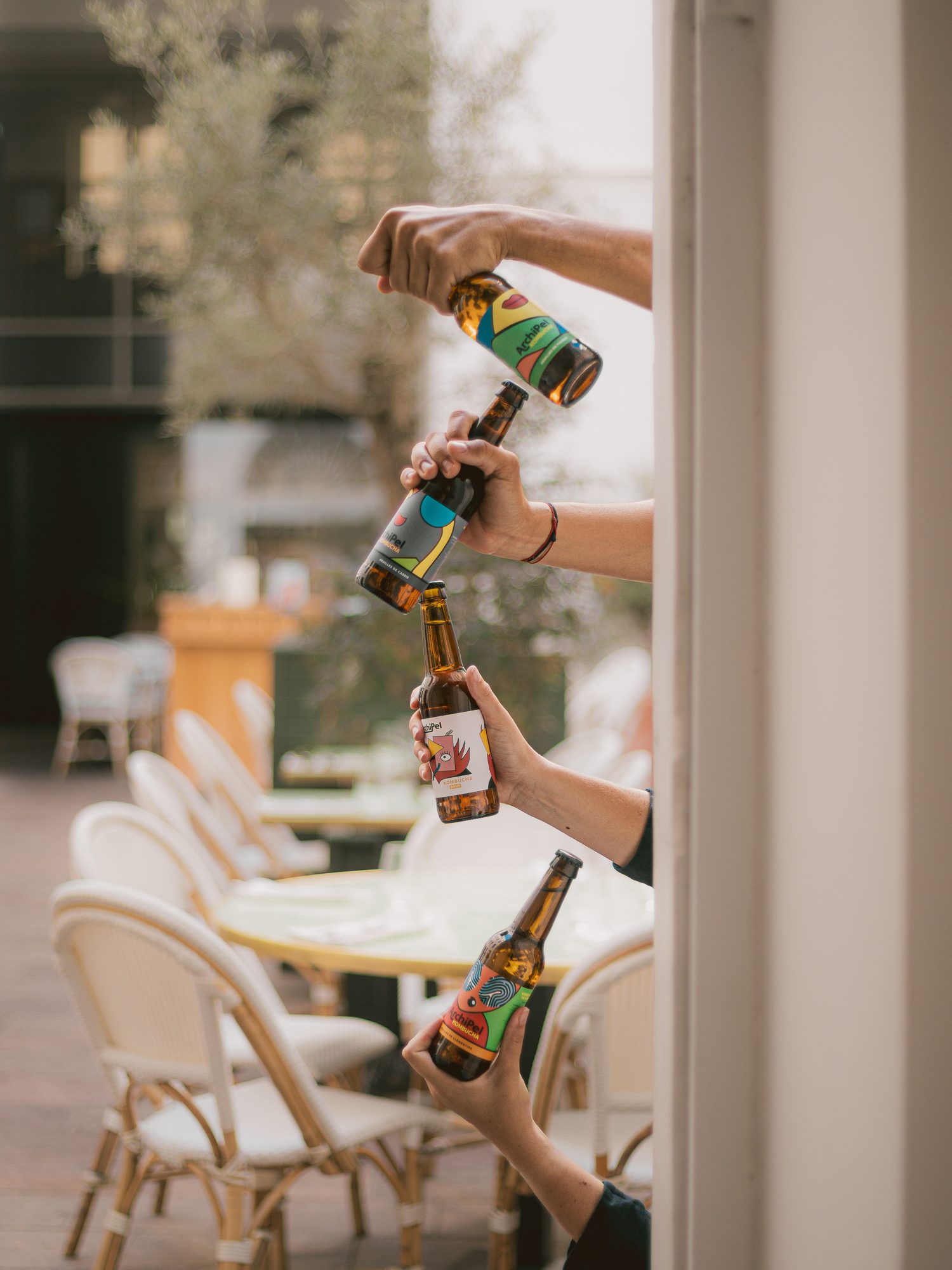 Quatre mains qui tendent des bouteilles de kombucha Archipel depuis une fenêtre