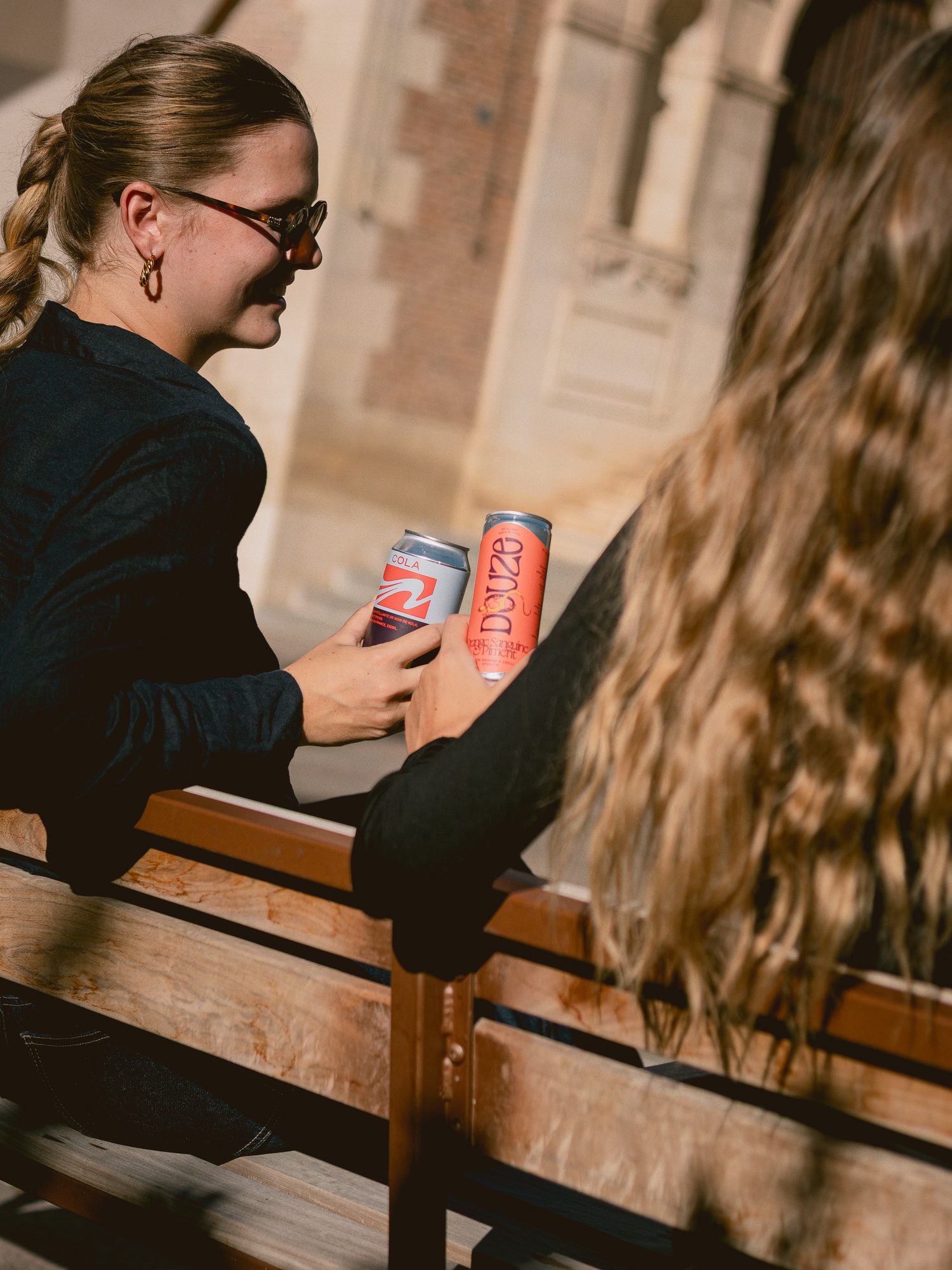 Deux amies trinquent sur un banc avec un cola UMÀ et un Douze orange sanguine et piment