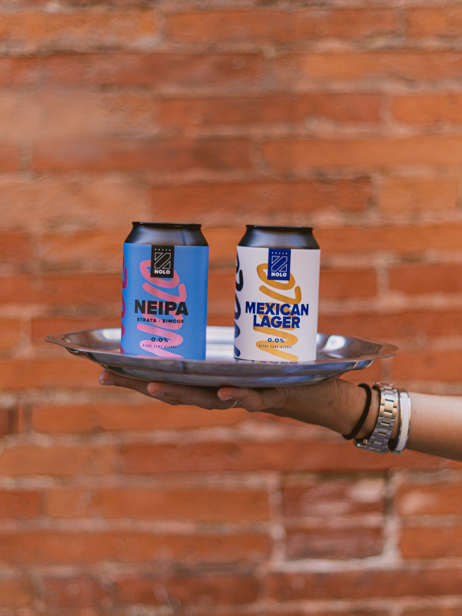 Deux canettes Prizm Nolo, NEIPA et Mexican Lager 0,0 %, sur un plateau devant un mur de briques
