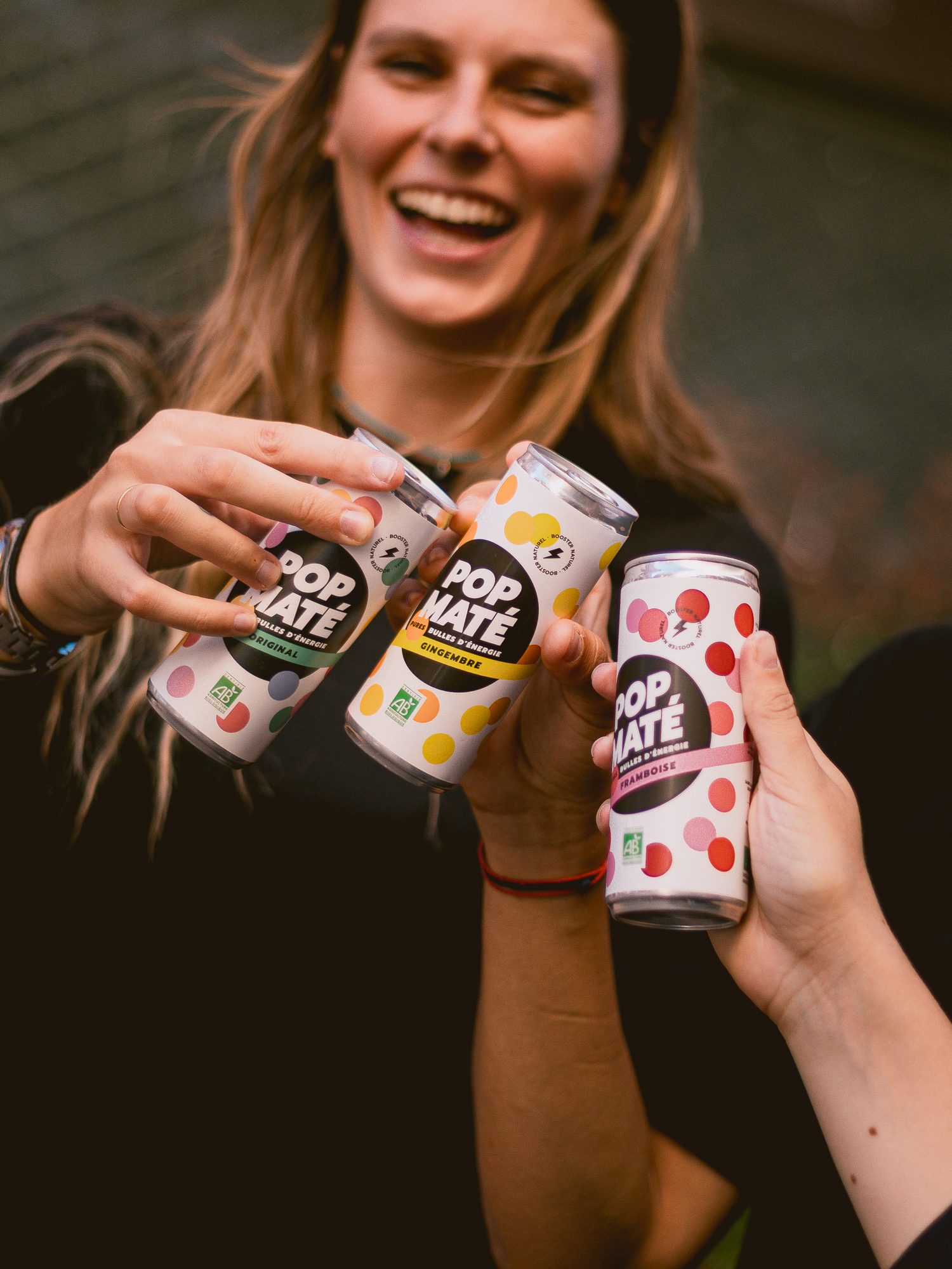 Trois canettes Pop Maté qui trinquent devant une amie qui rit